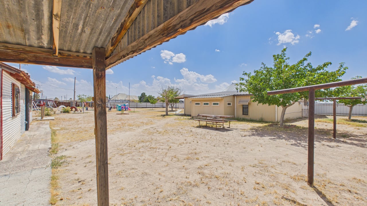 Fenced backyard with a covered wooden patio, shade tree, picnic table, and detached outbuilding.