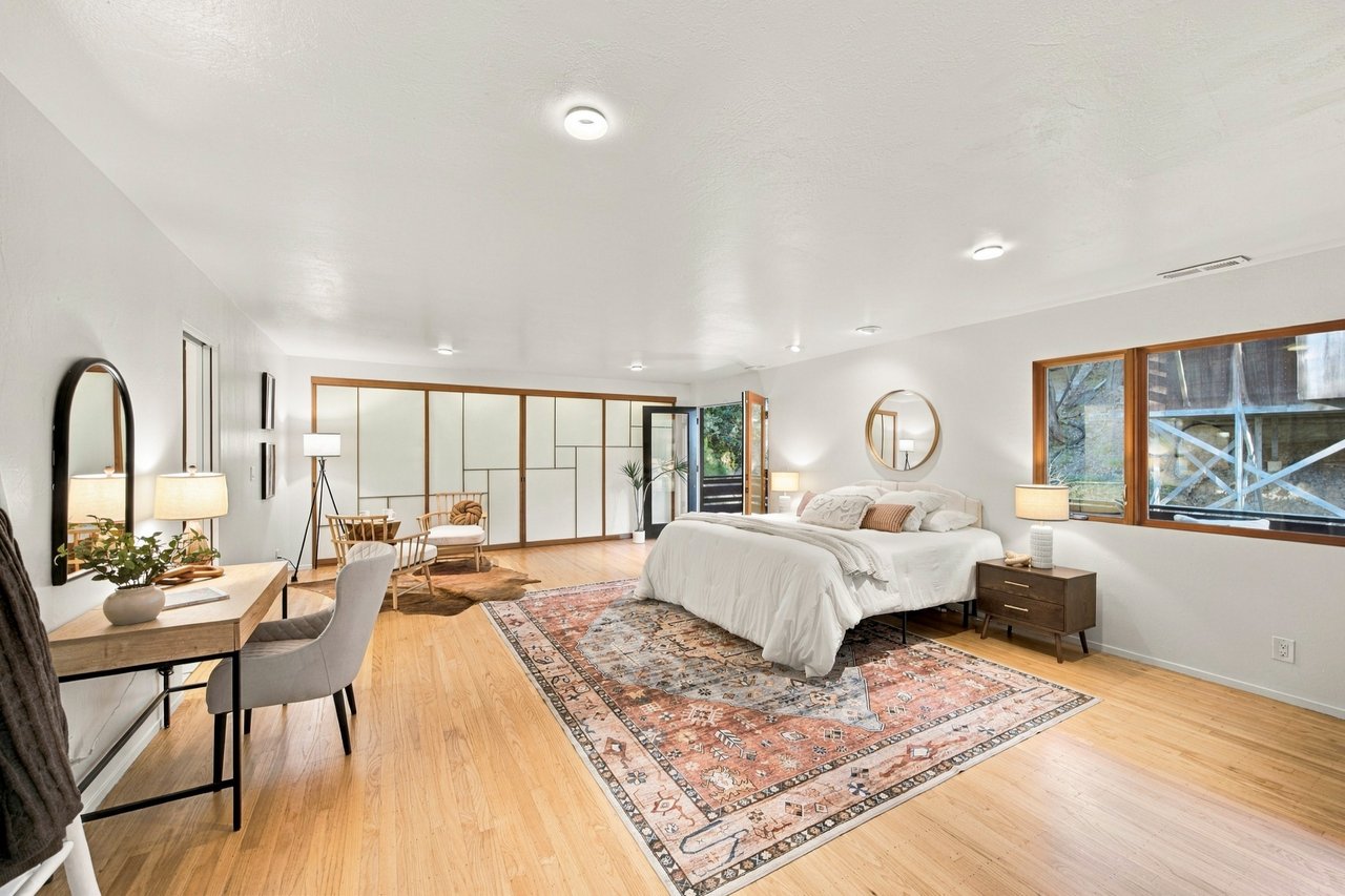 Staged bedroom with a large bed, hardwood floors, patterned rugs, and wide windows.