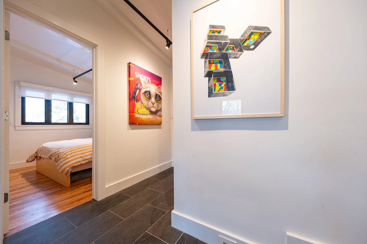 Renovated hallway opening into a bedroom with hardwood floors, a striped bedspread, and colorful framed artwork.