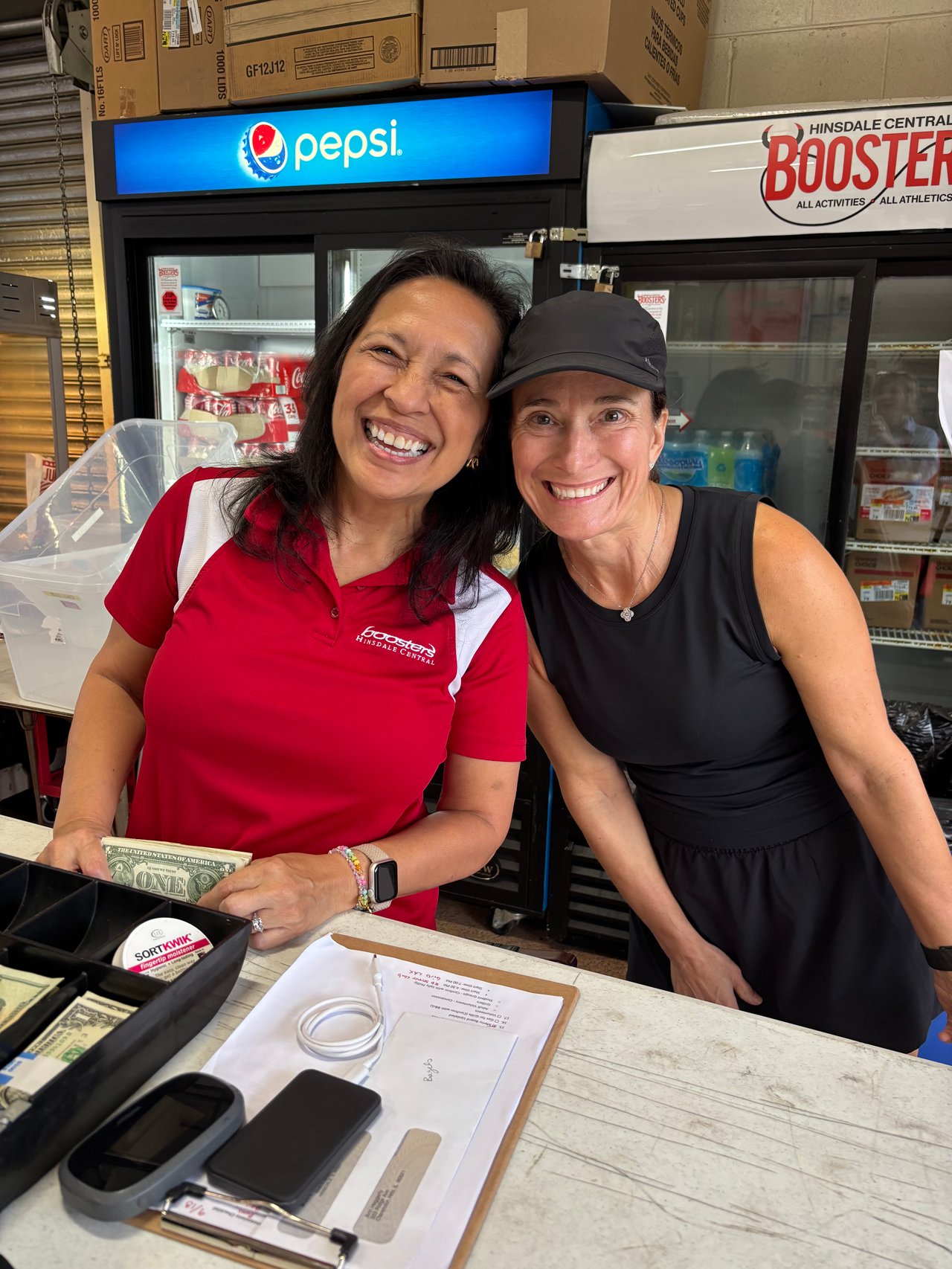 Two women smile at the Hinsdale Central Boosters counter, with a Pepsi sign and concession refrigerators behind them.