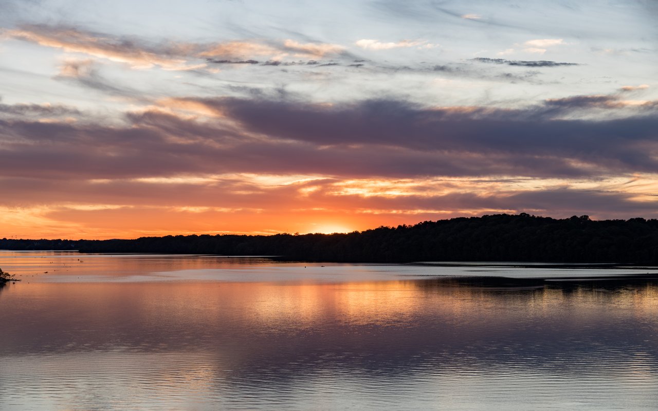 Calm lake reflecting an orange sunset beneath layered clouds and a wooded shoreline.