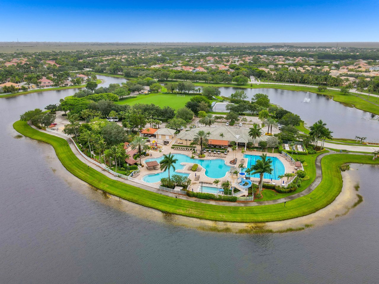 Aerial view of a waterfront community with pools, clubhouse, tennis court, and palm-lined paths.