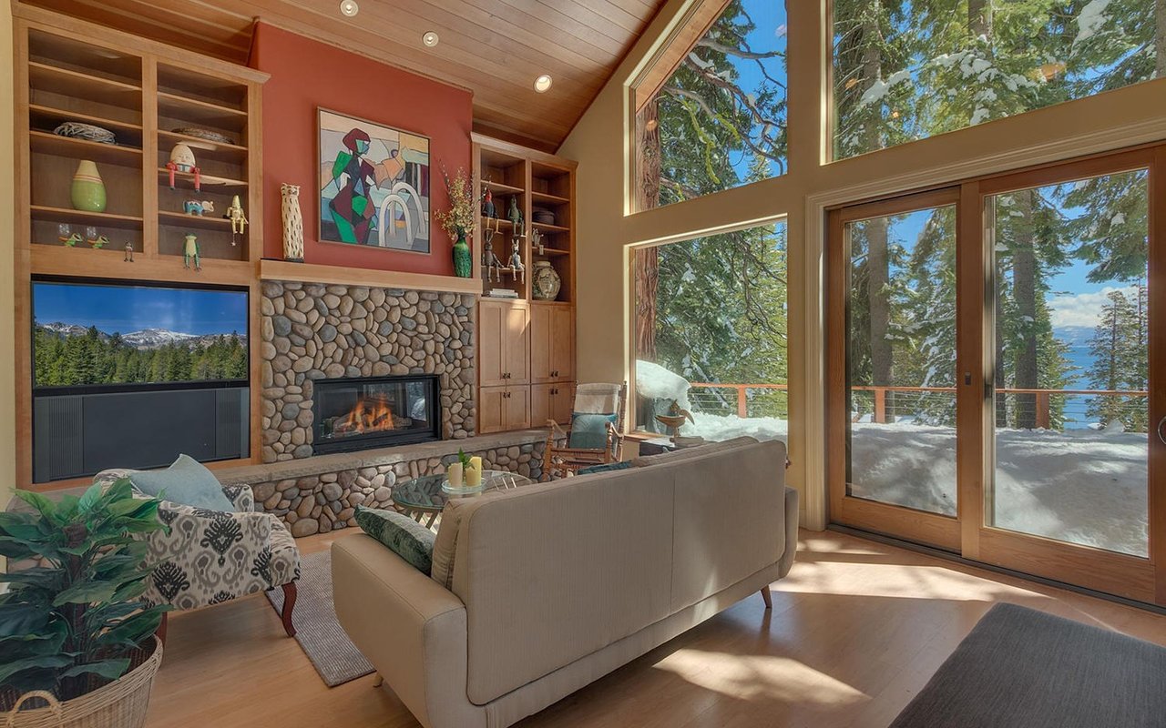 Living room with a stone fireplace and large windows overlooking snowy evergreens and a lake.