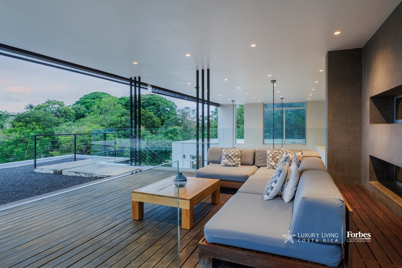 Modern living room with a large sectional sofa, wooden deck, and views of dense green trees.