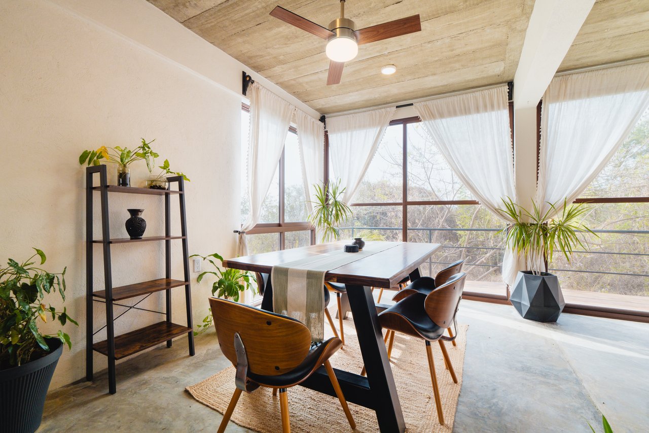 Dining room with a dark wood table, ceiling fan, and large curtained windows overlooking trees.
