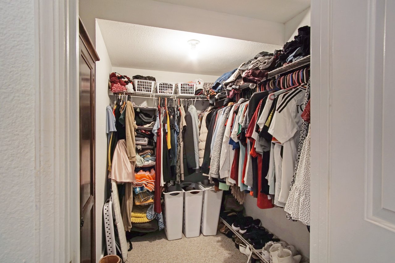 A well-organized walk-in closet with clothes neatly hung on racks, folded items on shelves, and white laundry bins below. Bright lighting creates an orderly, inviting space.