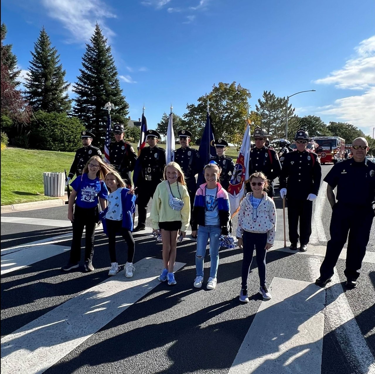 Uniformed parade participants and girls carrying flags cross a street in a sunny parade procession.