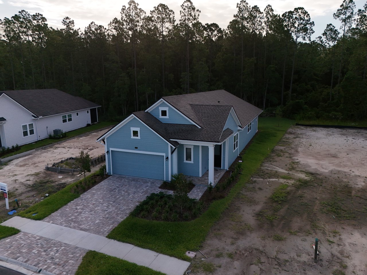Blue house with a paved driveway and partial landscaping, surrounded by sandy lots and pine trees.