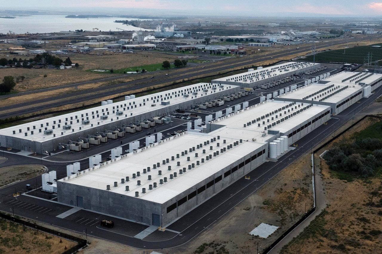 Aerial view of a large, modern data centre complex with cooling units, set in a semi-industrial area near a highway.