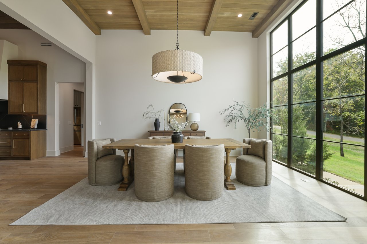 Dining room with a wood-beamed ceiling, upholstered chairs, and tall windows overlooking greenery.
