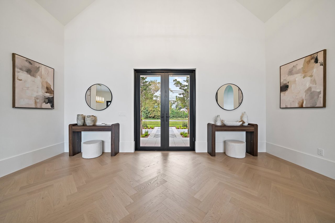 High-ceilinged foyer with herringbone floors and black-framed glass doors opening to a landscaped walkway.