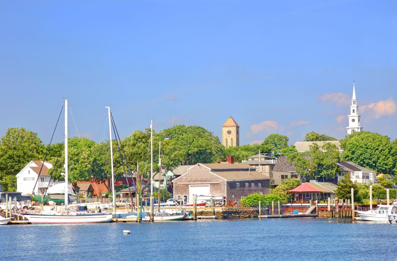 Harbor village with moored sailboats, waterfront homes, and church towers.