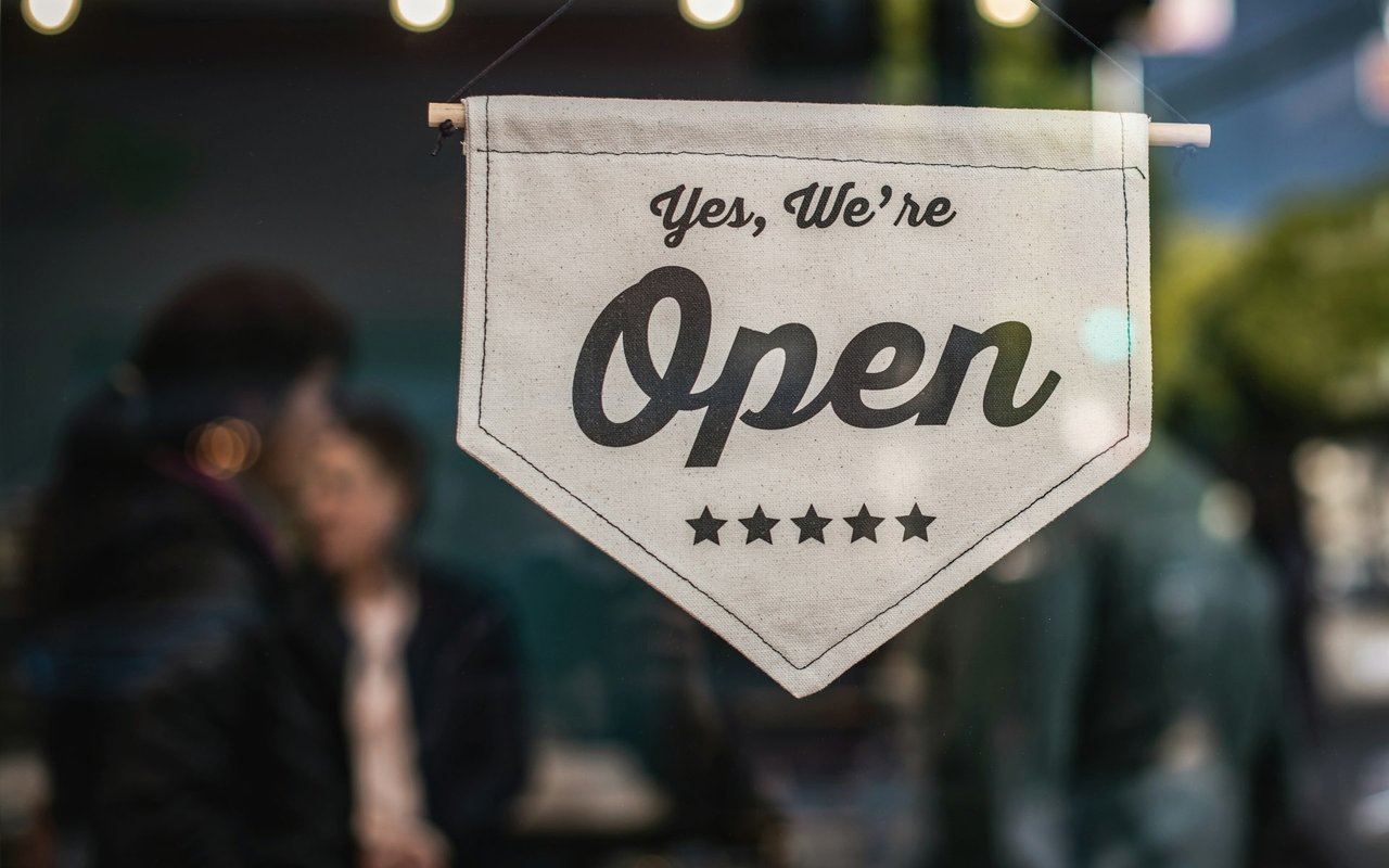 An open sign hangs in a shop window, with people blurred behind it.