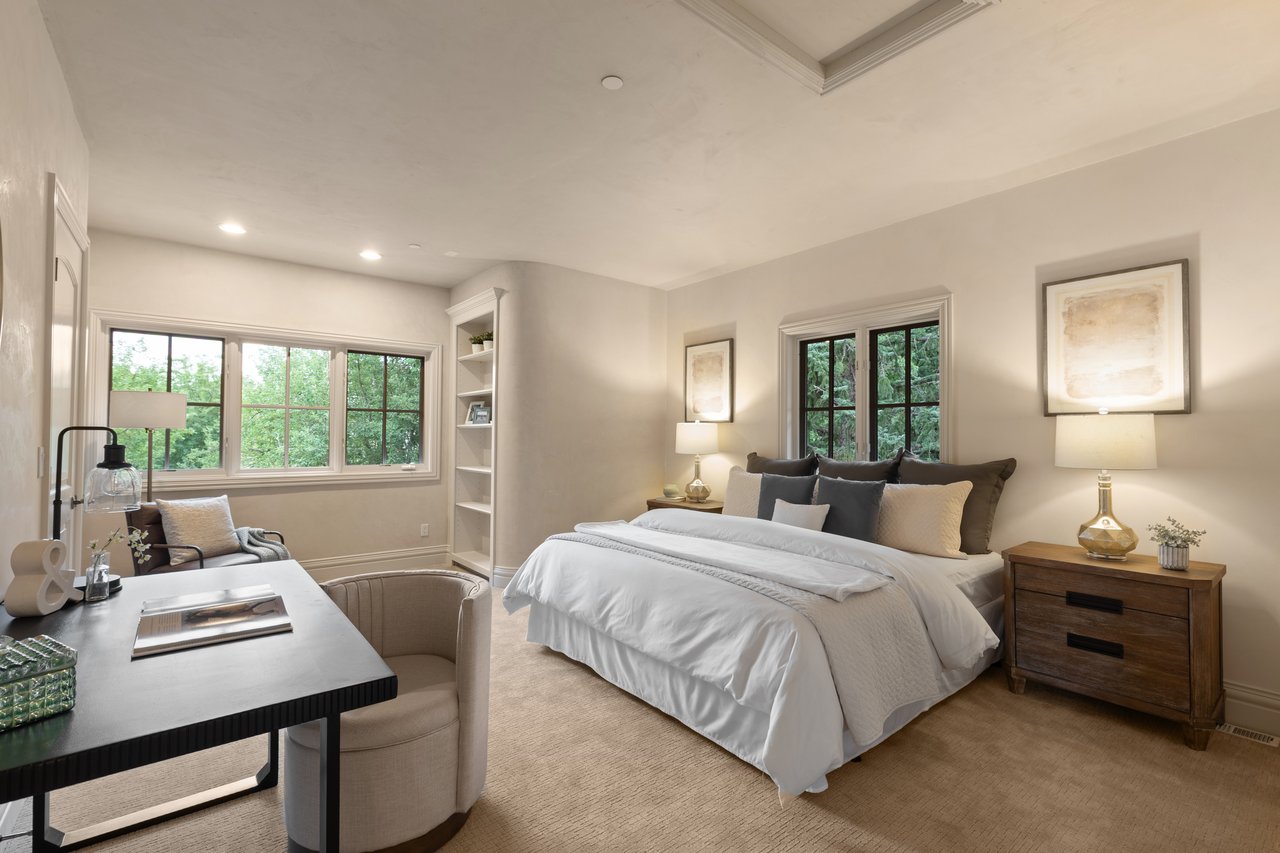 Bedroom with a white-dressed bed, wood nightstands, and large windows overlooking leafy trees.
