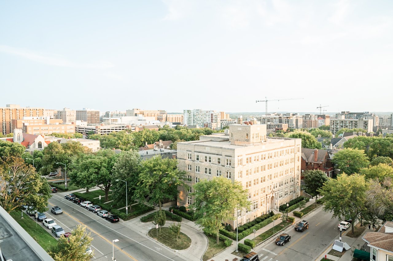 An aerial view of downtown Madison, Wisconsin, showing tree-lined residential streets, traffic, and the city skyline in the background, representing the mix of urban and residential corridors that shape the Madison commute experience.