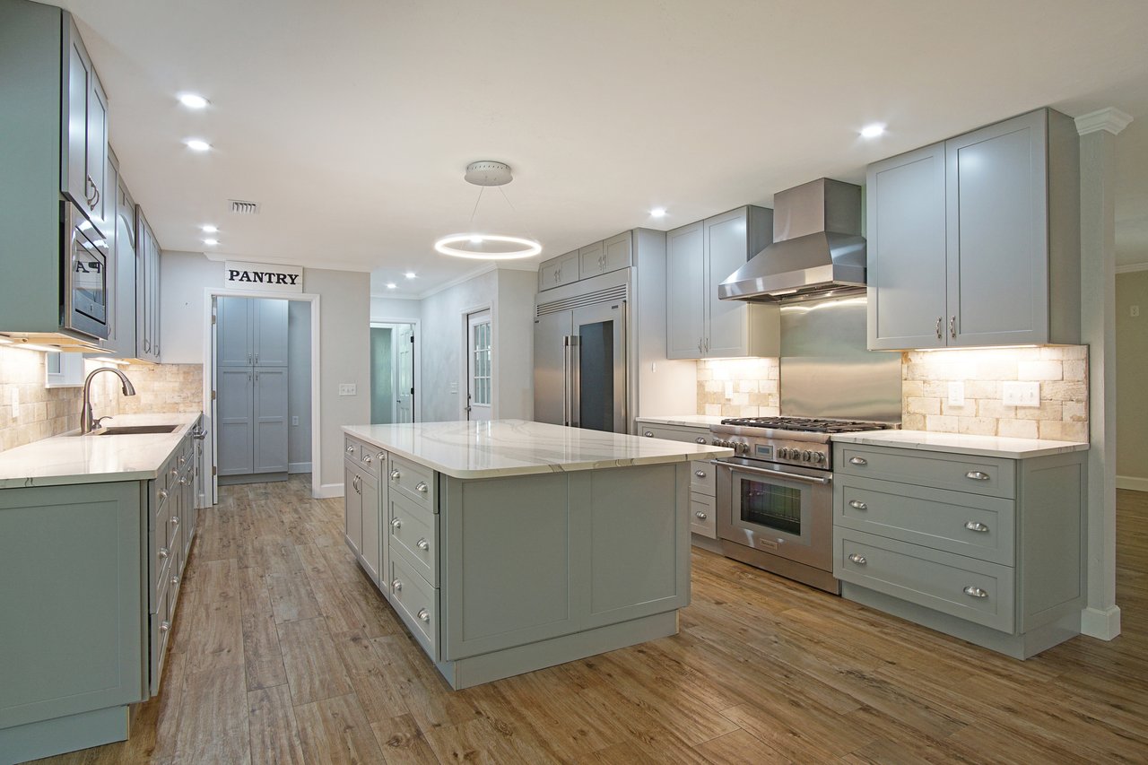 Modern kitchen with gray cabinets, a large island, stainless steel appliances, and wood flooring. Bright lighting creates an inviting atmosphere.