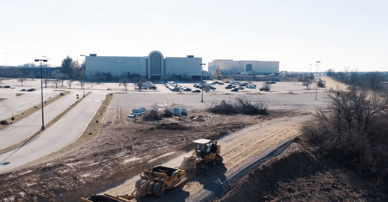 Site grading at the former Chesterfield Mall, with heavy excavators moving dirt across the redevelopment site and the remaining Dillard’s building in the background.