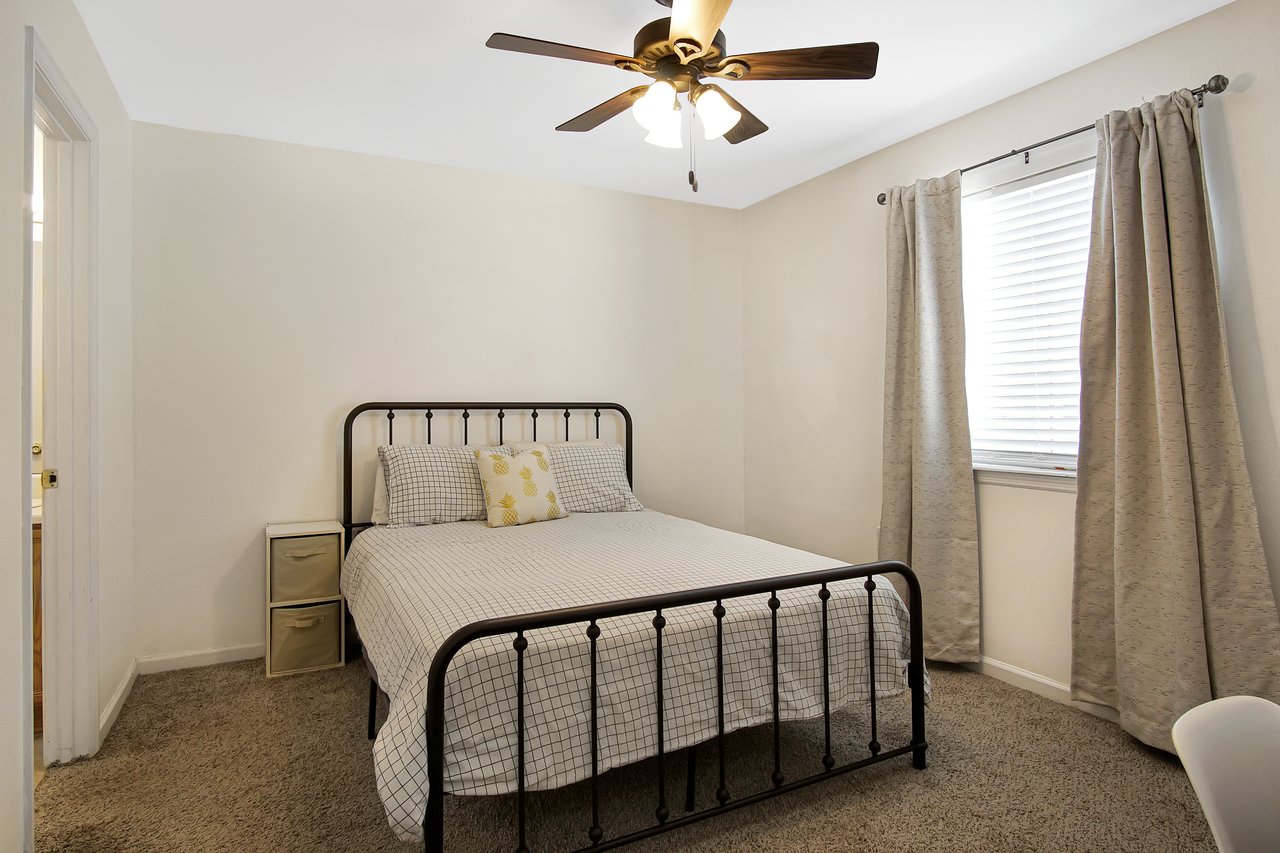 Simple bedroom with a black metal bedframe, checkered bedding, and a yellow accent pillow. Beige curtains hang beside a window with blinds. Ceiling fan above.