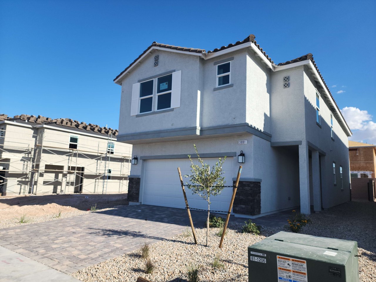 Two-story gray stucco home with a white garage, stone accents, and a newly planted tree in front.