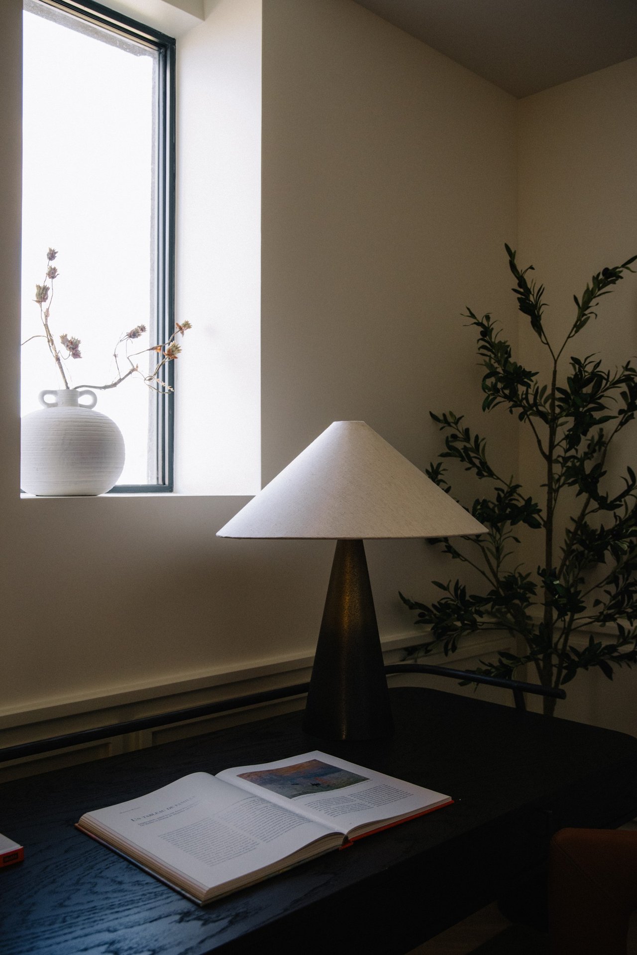 Interior vignette with a sculptural lamp, open book, leafy plants, and a bright window.