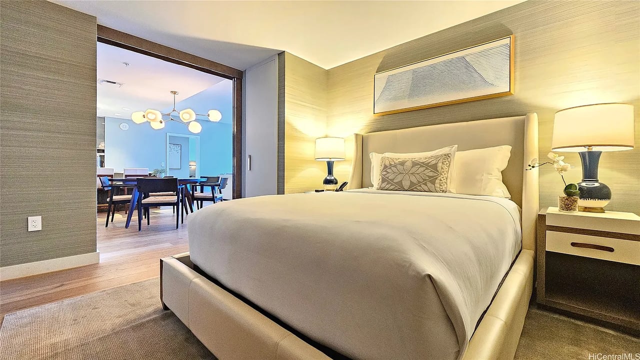 Bedroom with an upholstered bed and bedside lamps, opening onto a dining area.