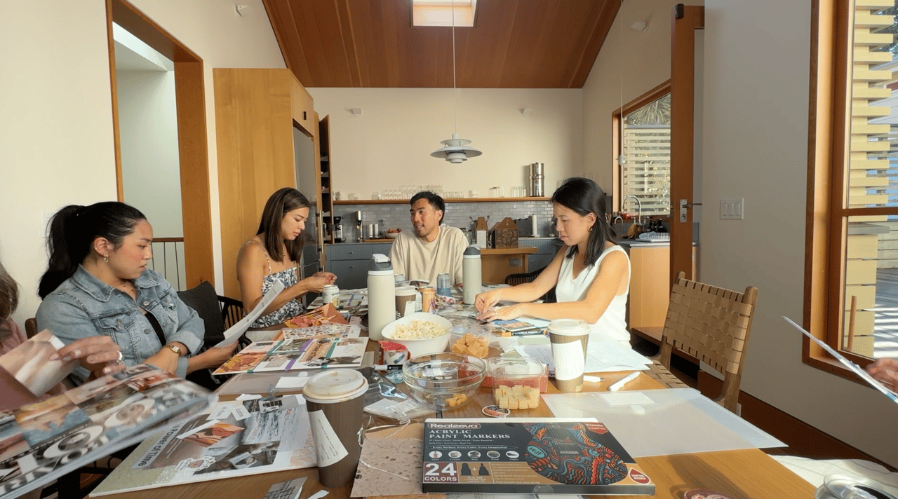 A group gathers around a kitchen table with papers, art supplies, and snacks.