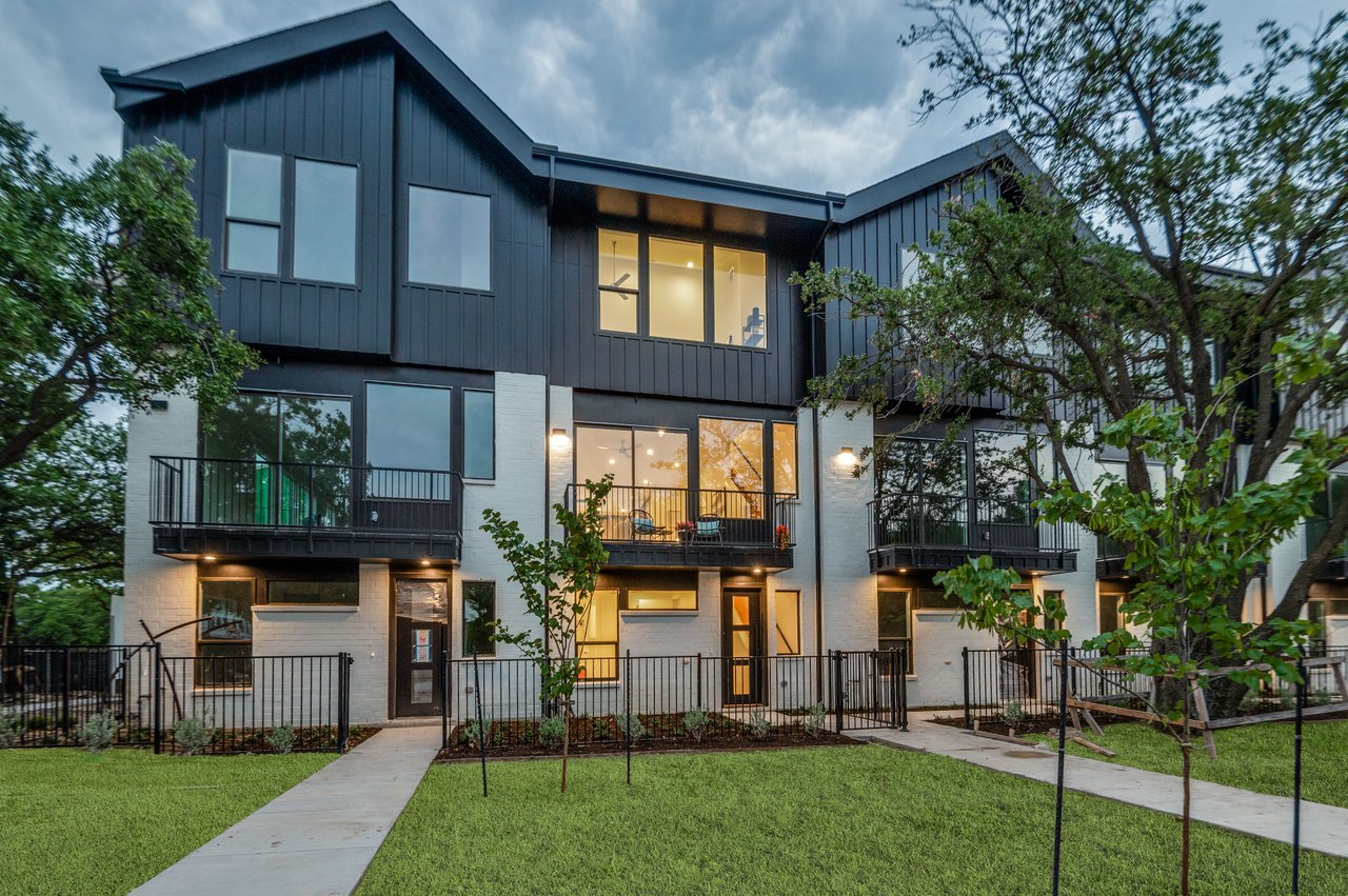 Modern townhomes with dark siding, white brick, balconies, and landscaped front yards at dusk.