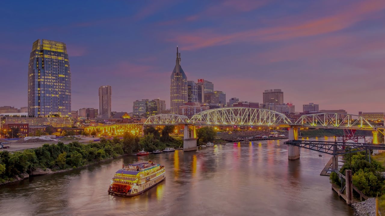 Nashville skyline at dusk across the Cumberland River, with a boat and steel truss bridge.