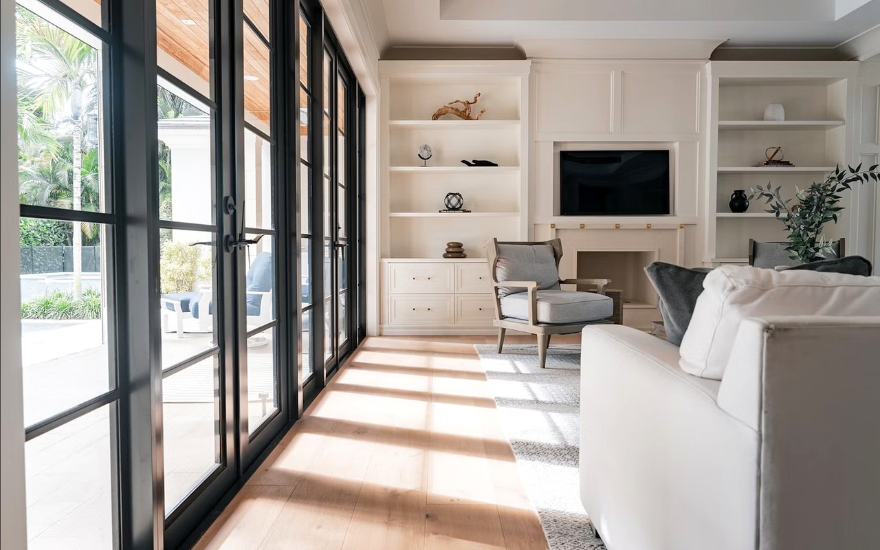 Bright living room with white built-ins and fireplace beside black-framed glass doors opening to a palm-lined patio.