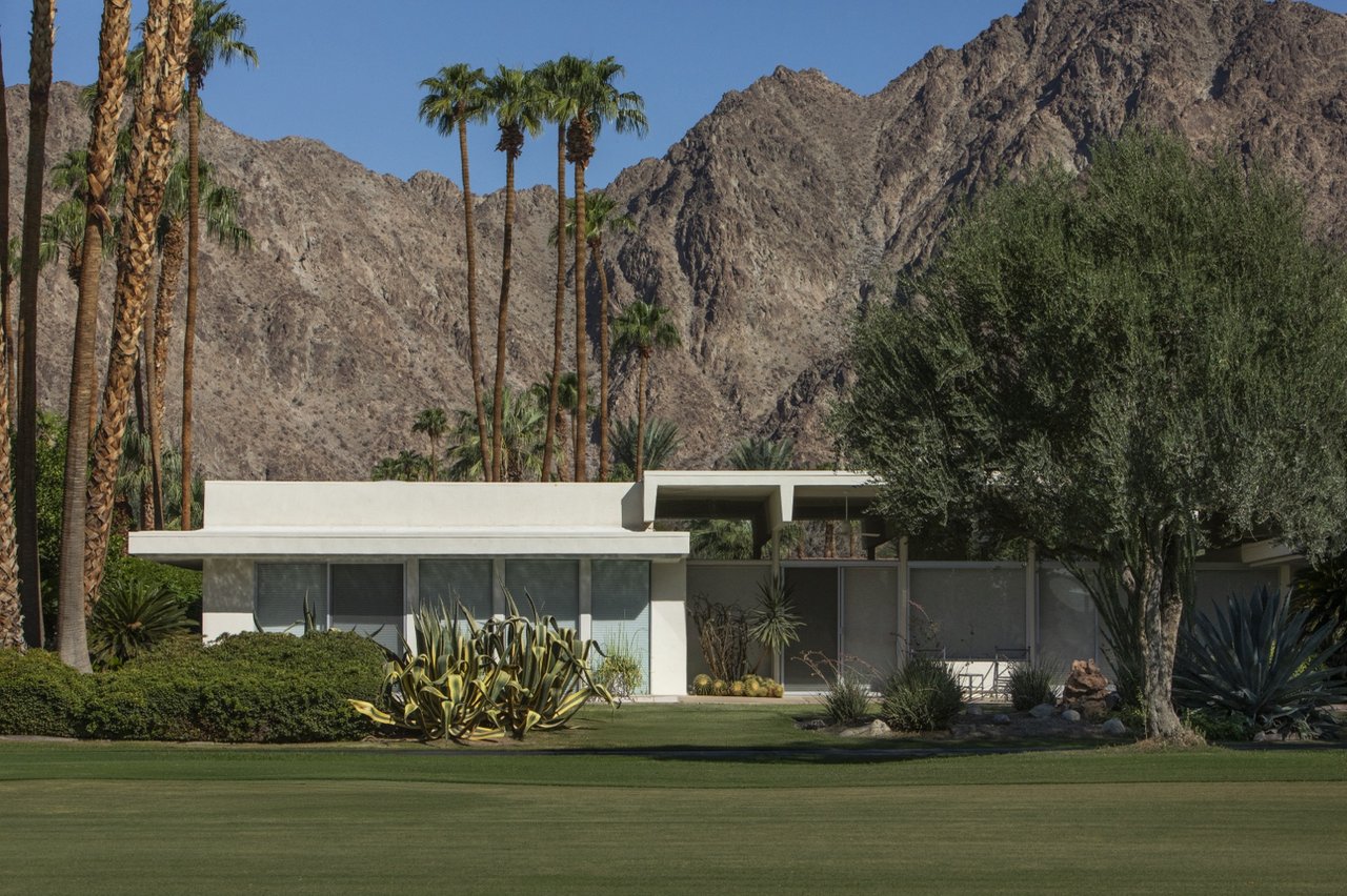 Modernist clubhouse beside a golf course, framed by palms and rugged desert mountains.