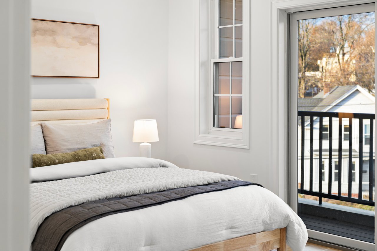 Bright bedroom with layered white bedding and a glass balcony door overlooking autumn trees.