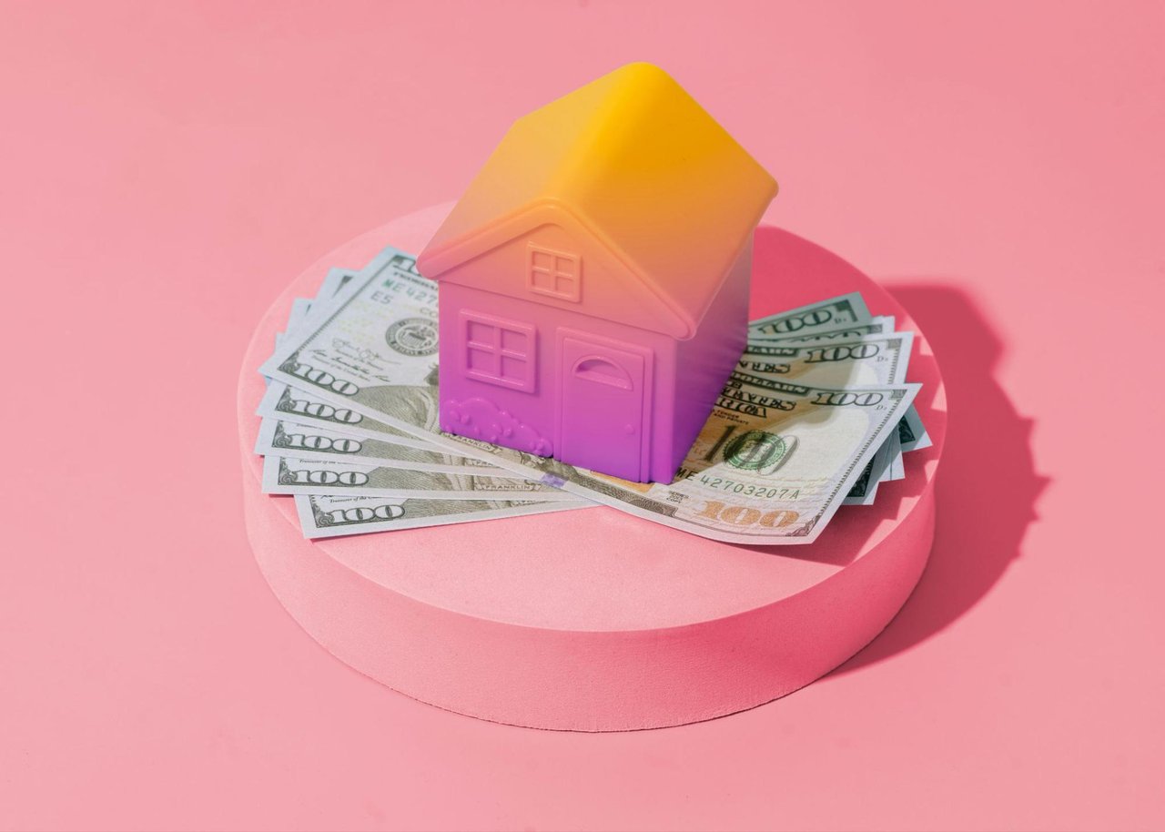 A colorful toy house sits atop scattered hundred-dollar bills on a pink platform.