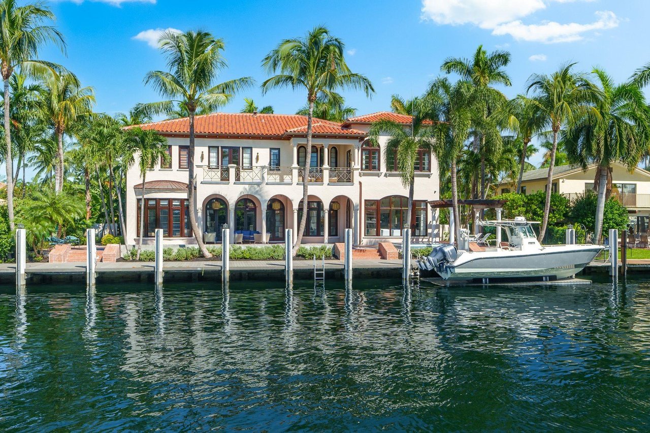 Two-story canal-front home with a terracotta roof, palm trees, and a motorboat at its dock.