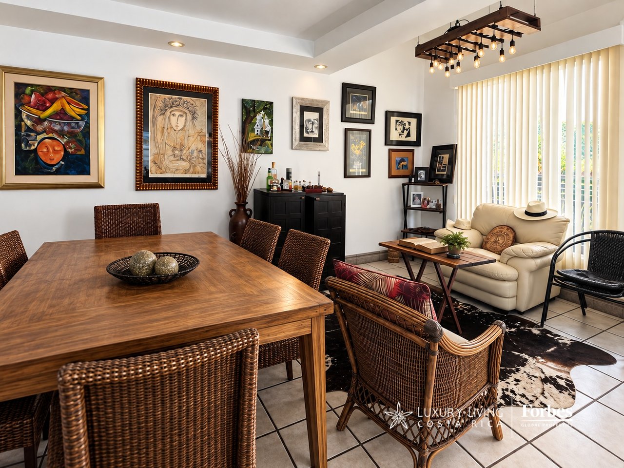 Furnished dining and living area with a large wooden table, woven chairs, cream sofa, and colorful framed artwork.