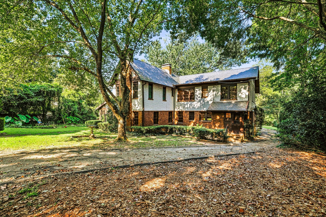 Two-story brick and stucco house with a slate roof, shaded by mature trees beside a leaf-covered driveway.