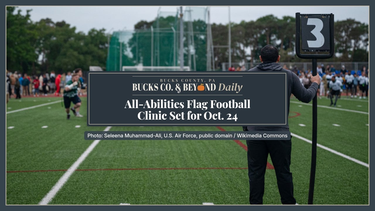 A sideline official, seen from behind, holds a number 3 down marker beside a green turf field during an adult flag football game, with players and spectators in the distance.