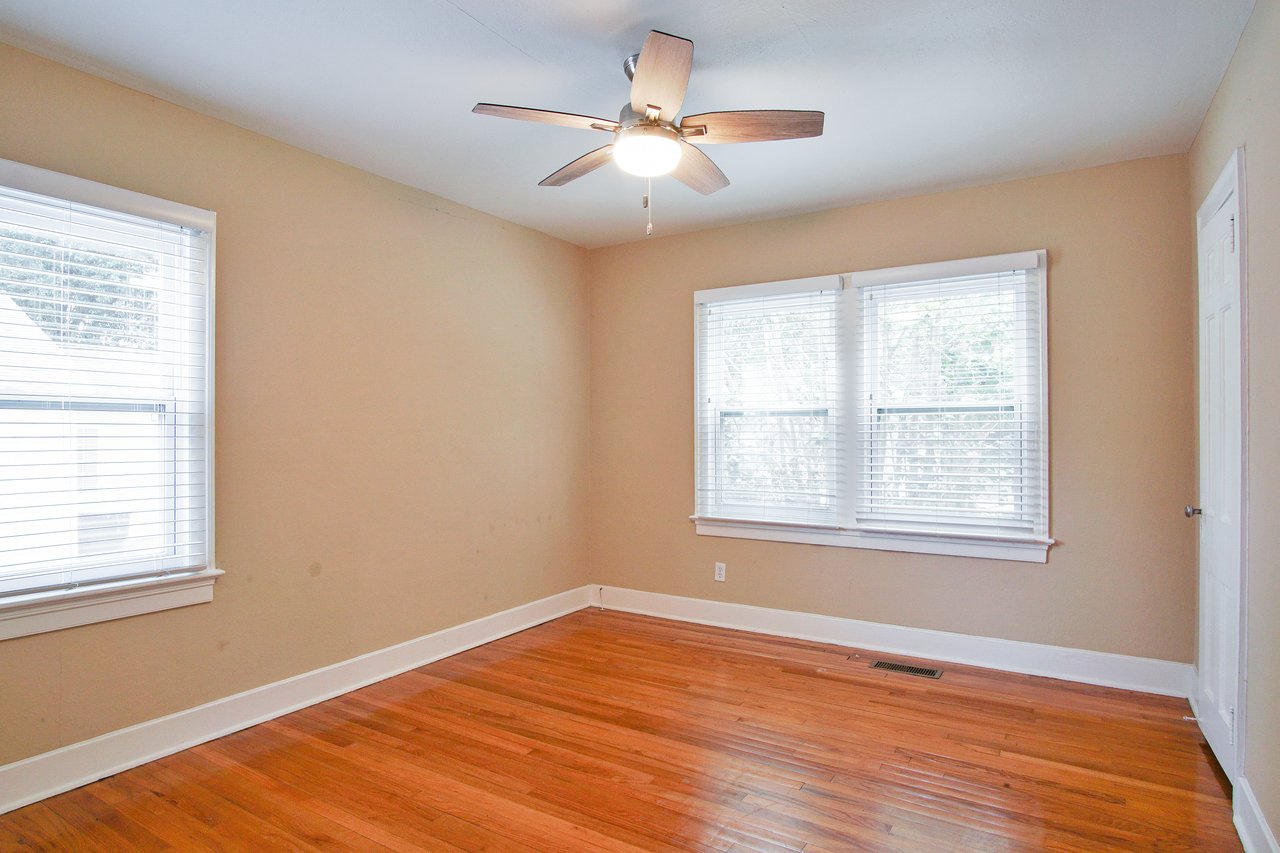 Empty room with light brown walls and wooden floor. Two windows with blinds allow natural light. A ceiling fan with a light fixture is centered above.