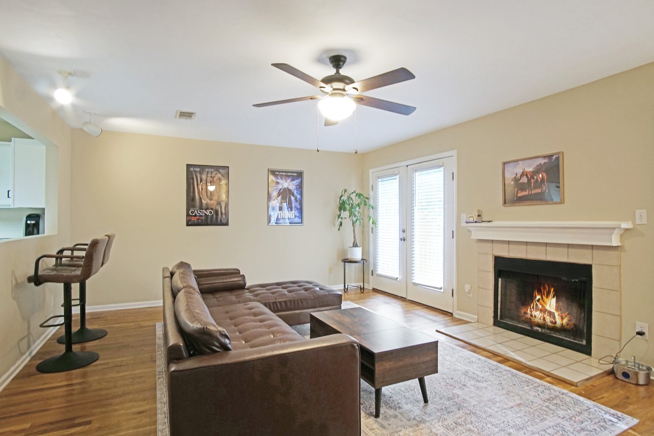 Living room at 529 Collinsford Road, Tallahassee, Florida, featuring a cozy fireplace with tile surround, French doors leading to the back deck, and hardwood flooring. The space includes a brown sectional sofa, coffee table, bar seating at the kitchen pass-through, and a ceiling fan with light fixture, creating a warm and inviting atmosphere.