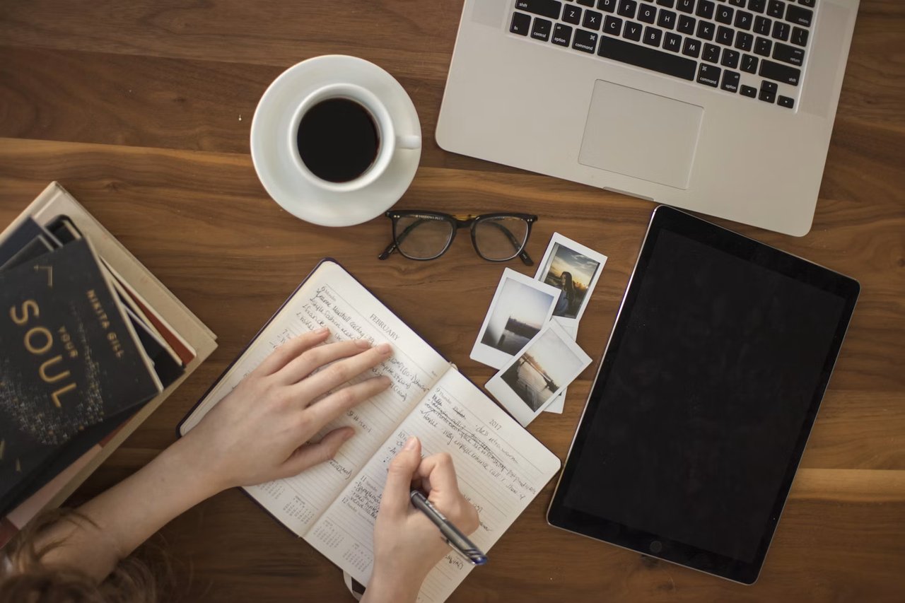 Hands writing in a journal beside coffee, a laptop, tablet, glasses, and photographs.