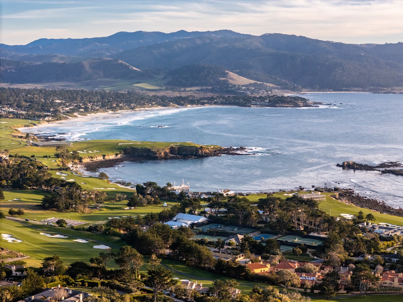 Aerial view of Pebble Beach golf course and coastline, with green fairways, tennis courts, and homes along a rocky shoreline backed by hills.