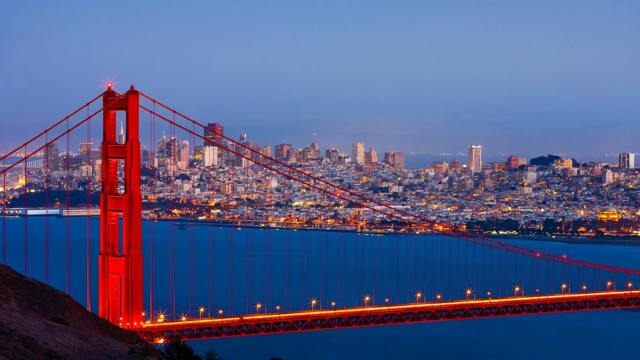 Evening view of Golden Gate Bridge with illuminated San Francisco skyline and hillside neighborhoods, showcasing the premium lifestyle and scenic beauty awaiting affluent individuals moving to San Francisco