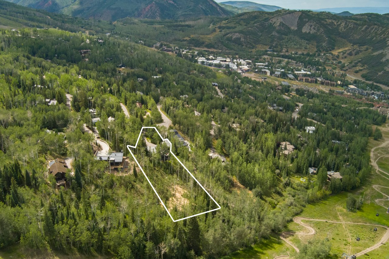 Aerial view of a wooded mountain parcel outlined in white, with homes and forested slopes nearby.