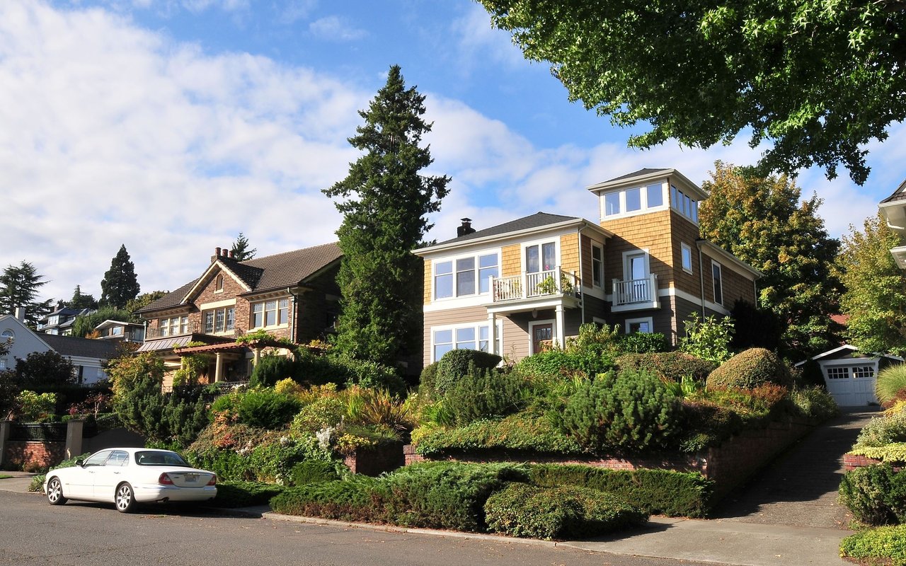 Two-story house with a shingled tower and landscaped slope among neighboring homes.