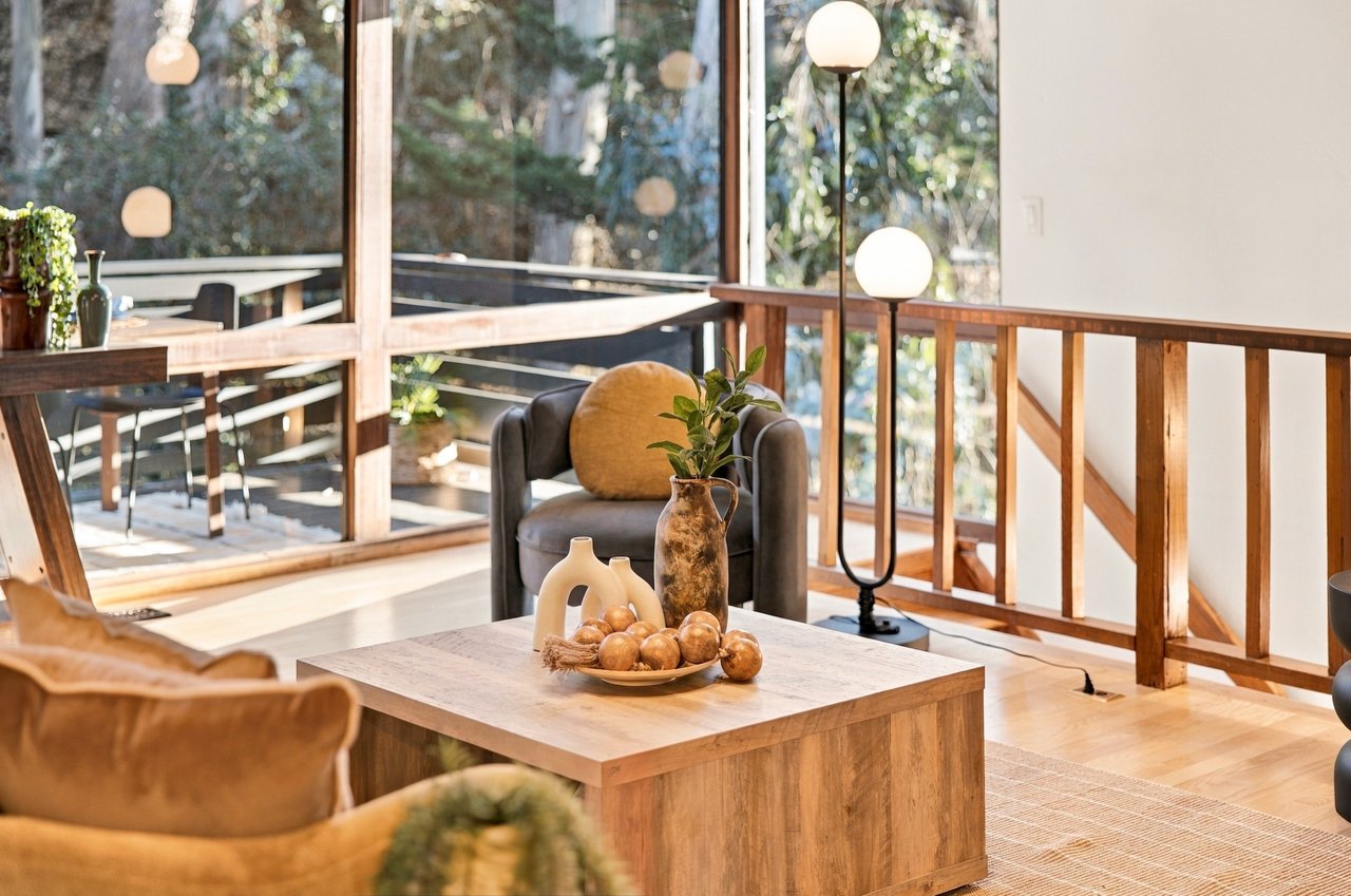 Staged living room with a gray armchair, wood coffee table, and floor-to-ceiling windows overlooking trees.