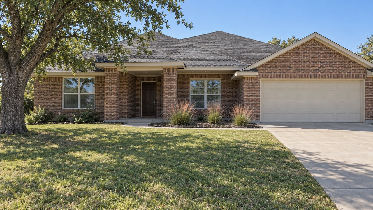 A single-story red-brown brick home with a deep front lawn, sheltered entry, concrete driveway and mature bur oak in bright daylight.