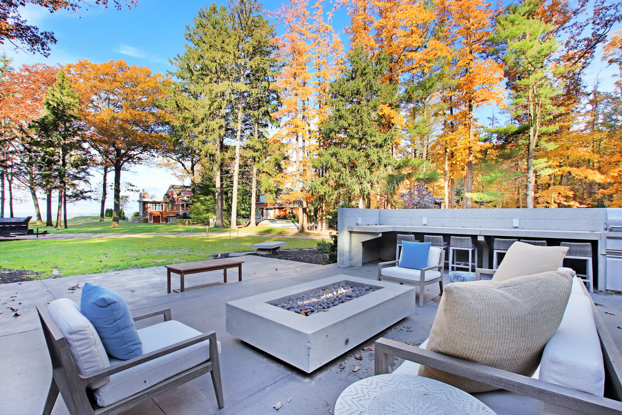 Lakeside patio with a concrete fire pit and lounge seating beneath autumn trees.