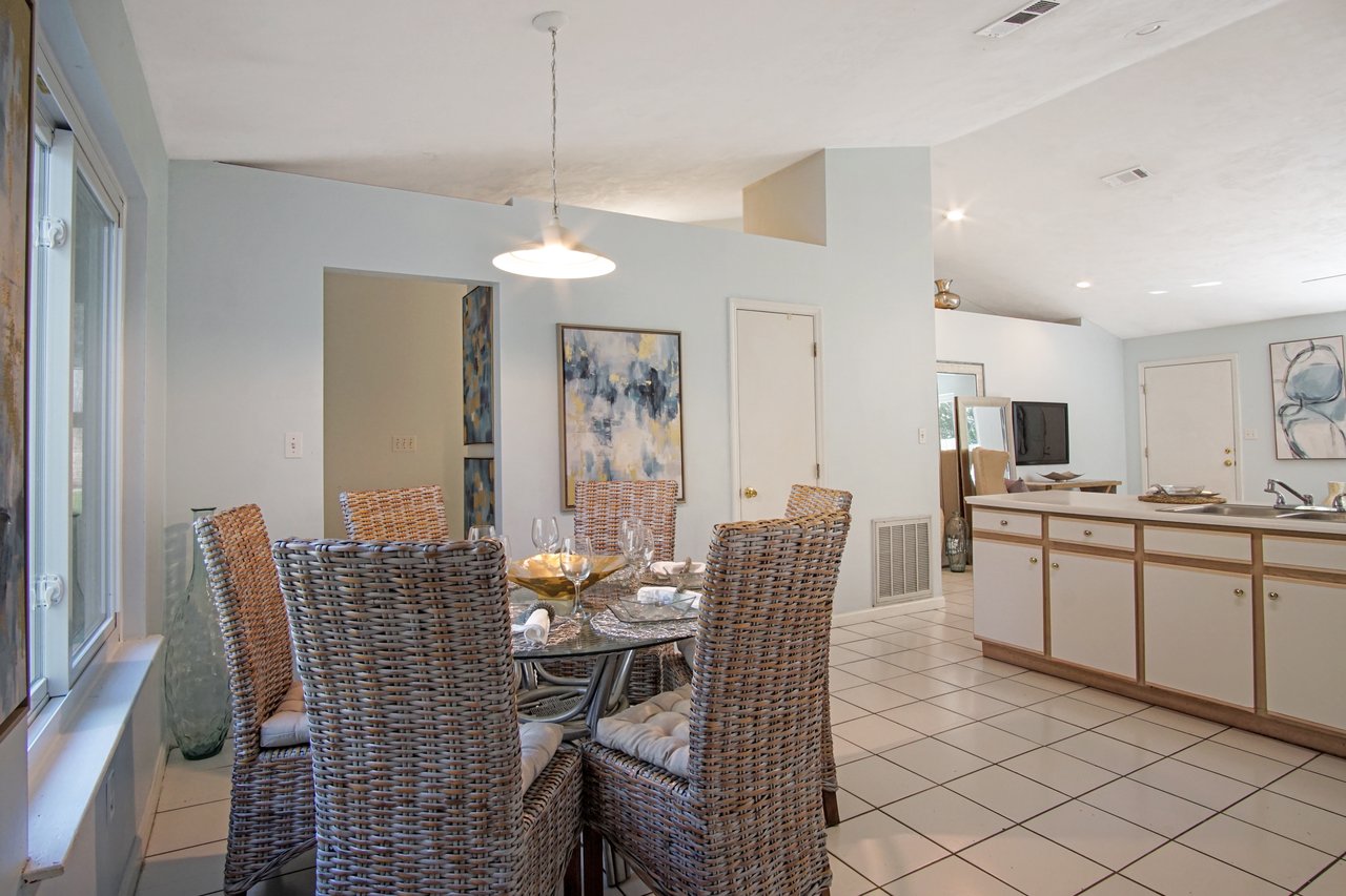 Dining room with wicker chairs around a glass table set with wine glasses and napkins. Soft lighting, modern abstract art, and light blue walls enhance the airy atmosphere.