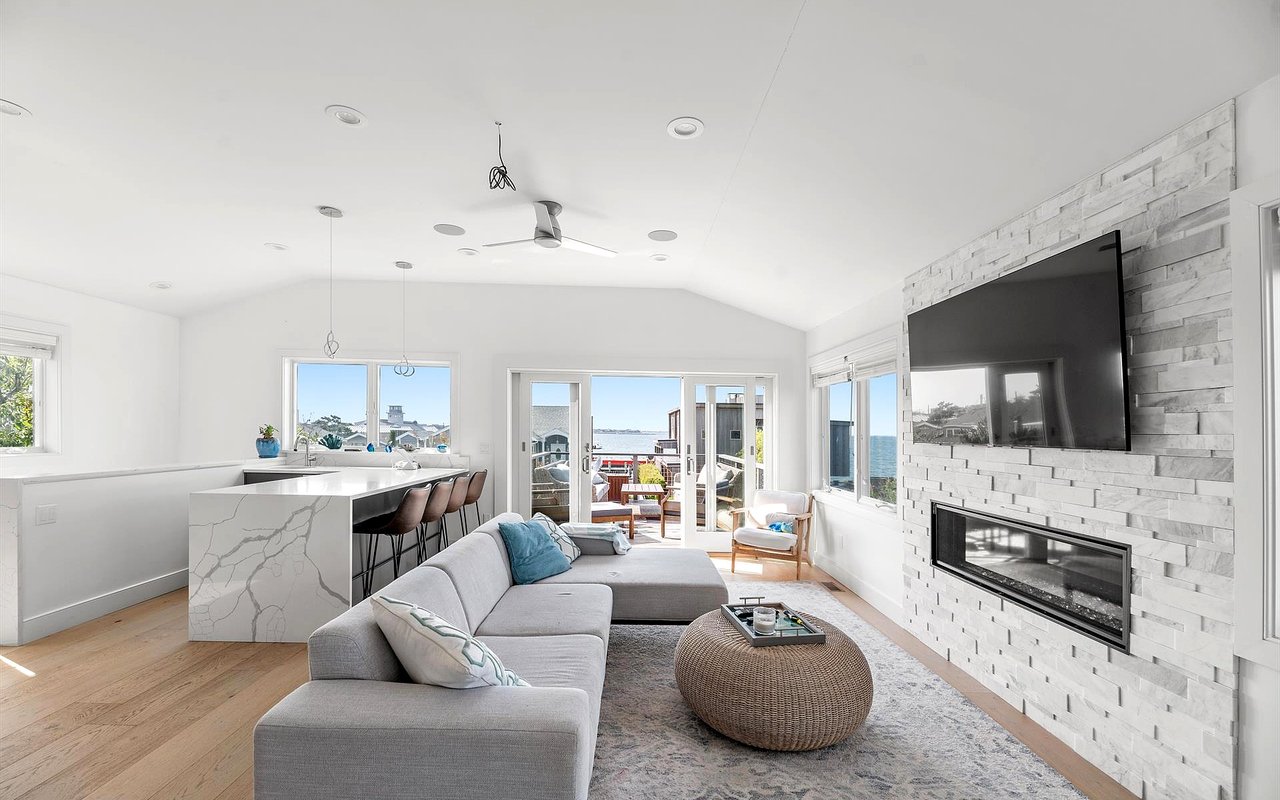 Open-plan living room with a gray sectional, stone fireplace, and windows overlooking the water.