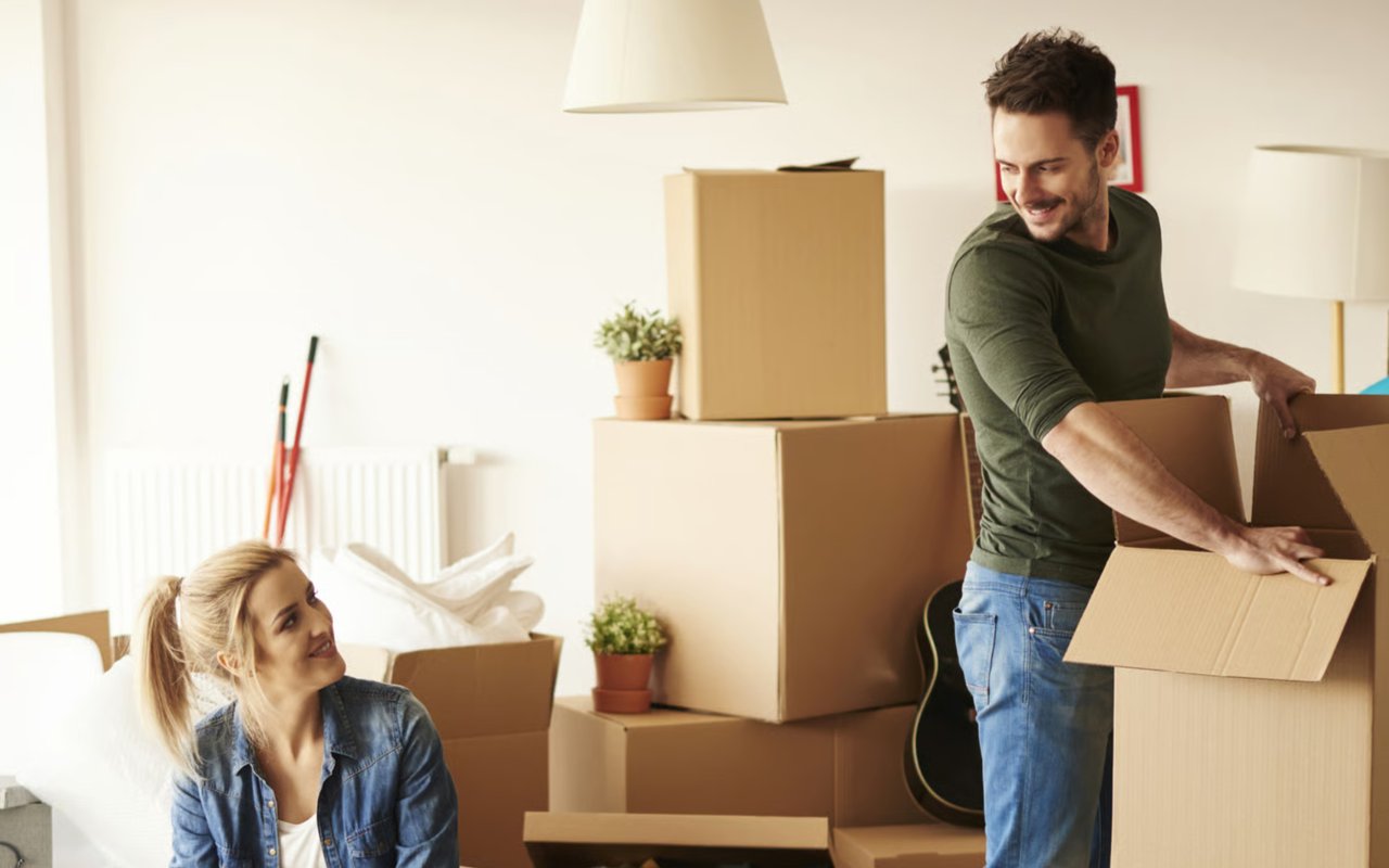 A happy couple unpacking moving boxes in their new home, with a woman looking at a man holding an open box.