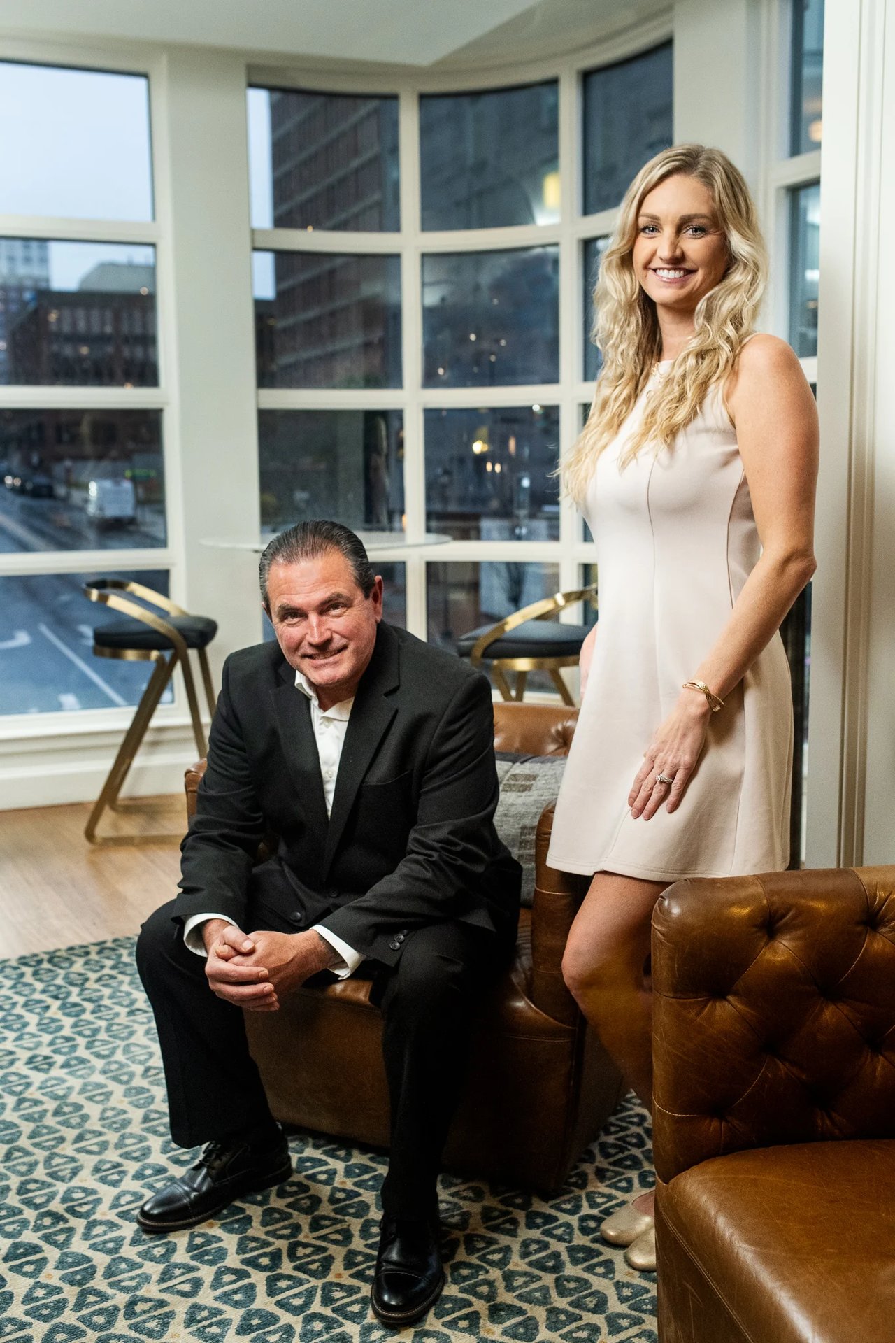 Two partners pose in an office, one seated in a black suit and the other standing in a white dress.